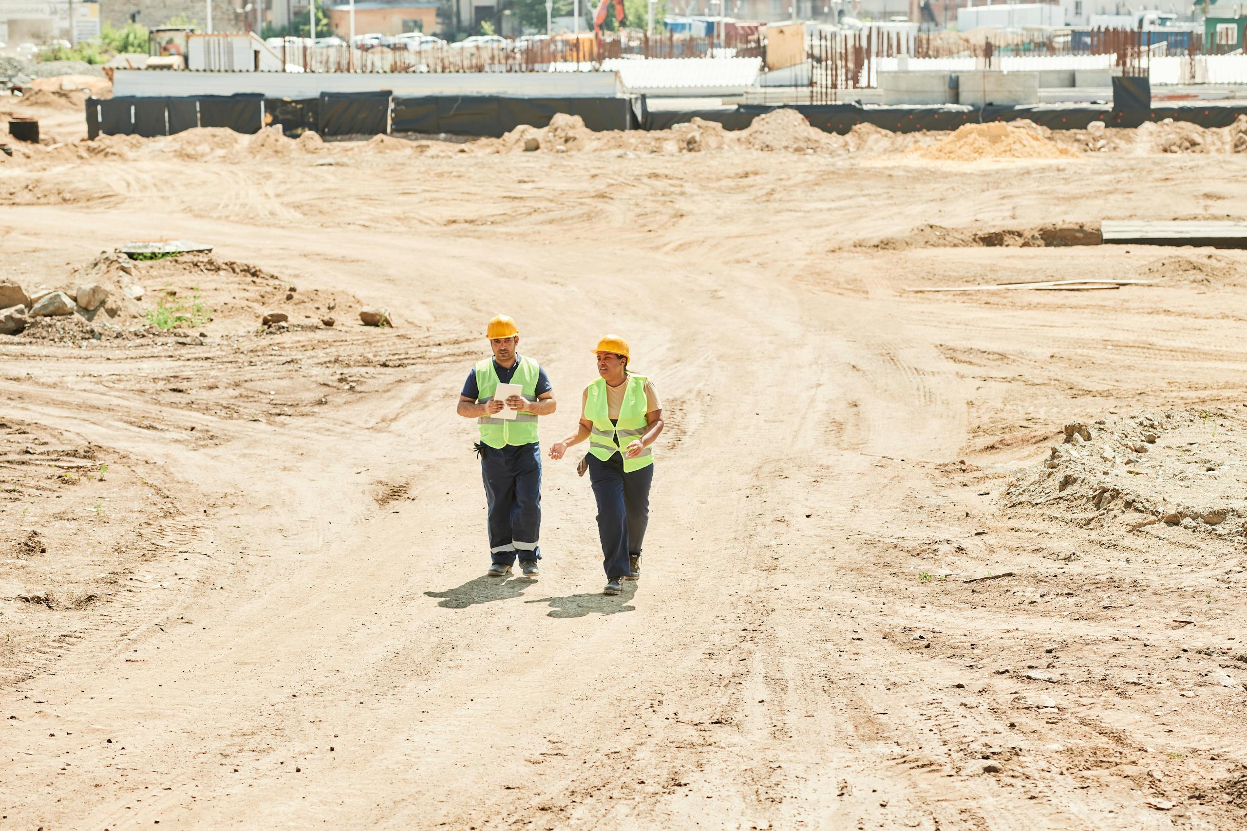 Two workers in safety gear walking across a construction site under a clear sky.